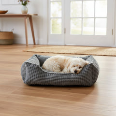 Kleiner weißer Hund schläft gemütlich in grauem Komfortbett Garik in einem hellen Raum mit Holzfußboden.