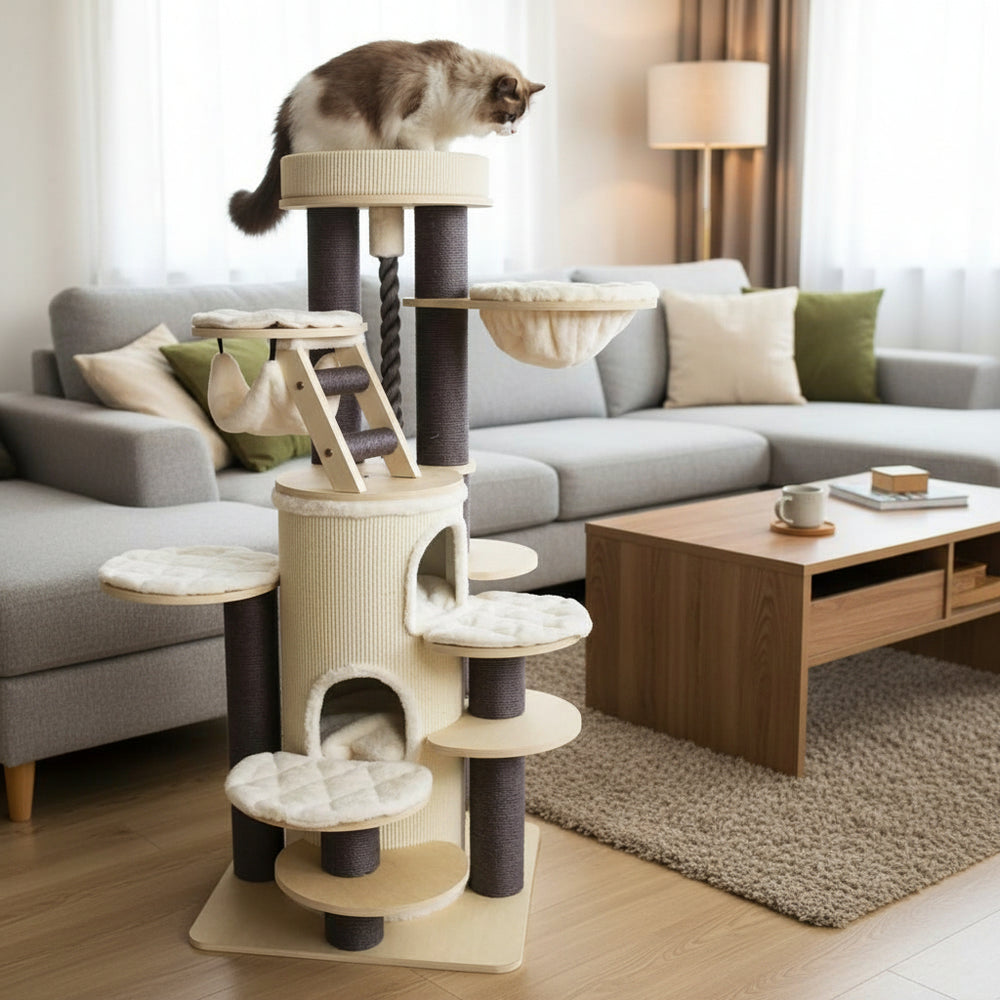 Beiger Kratzbaum CRIO mit mehreren Plattformen und einer sitzenden Katze im Wohnzimmer neben einem grauen Sofa.
