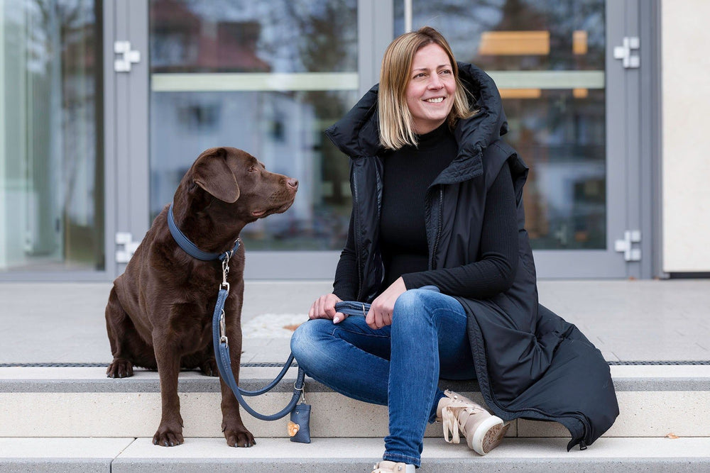 Frau mit schwarzem Parka sitzt auf Treppe neben braunem Labrador, lächelnd vor unscharfen Gebäuden.