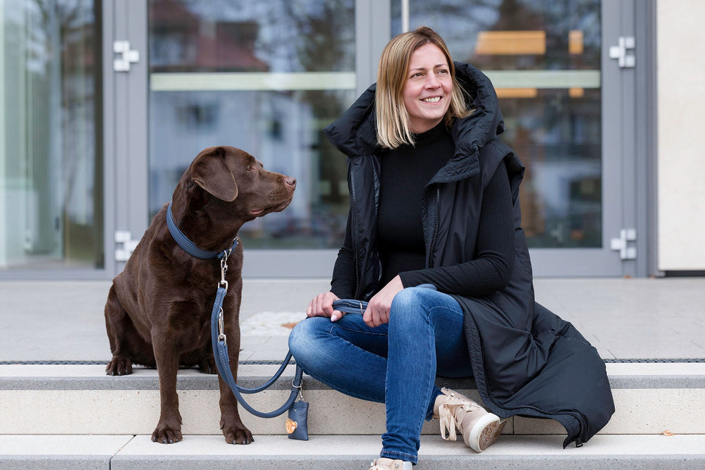 Frau mit schwarzem Parka sitzt auf Treppe neben braunem Labrador, lächelnd vor unscharfen Gebäuden.