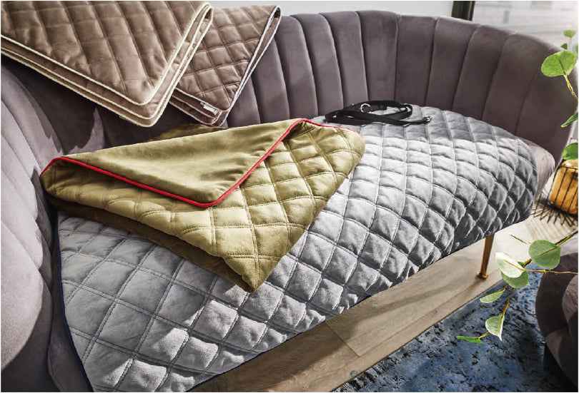 Moderne Couch mit grauem Hundedecken-Überwurf und weiteren in beige und olivgrün, dekoriert mit Pflanzen im Hintergrund.