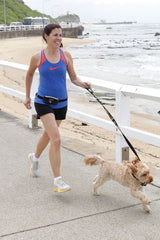 Frau joggt am Strand mit Hund, trägt DOOG Mini Gürteltasche, blaues Sporttop und schwarze Shorts unter sonnigem Himmel.