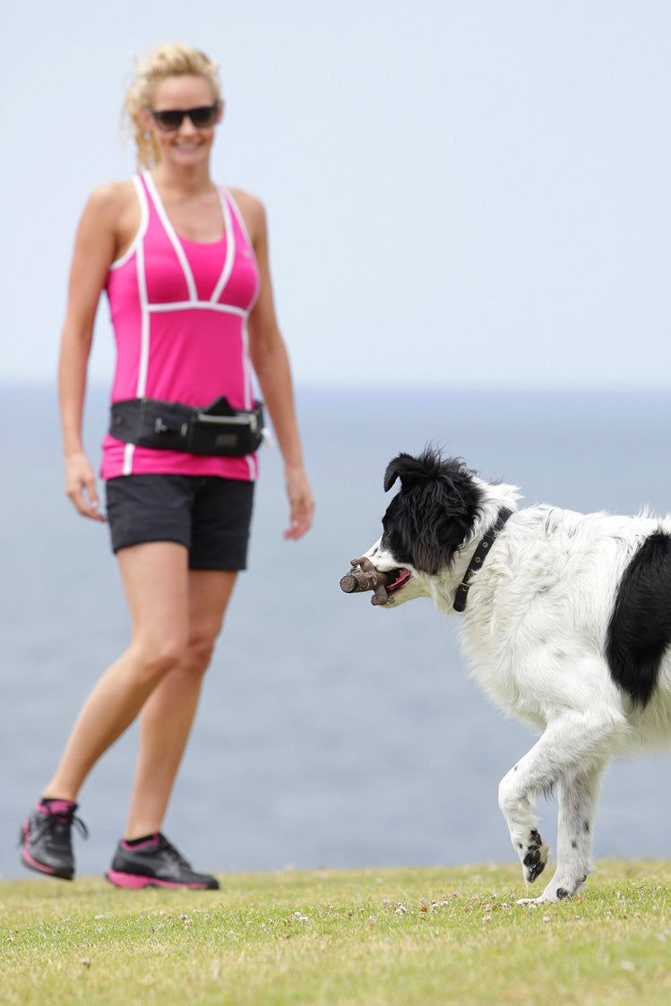 Frau in Sportkleidung beobachtet Hund mit Spielzeug auf Wiese vor blauem Himmel und Wasser. DOOG Gürteltasche sichtbar.