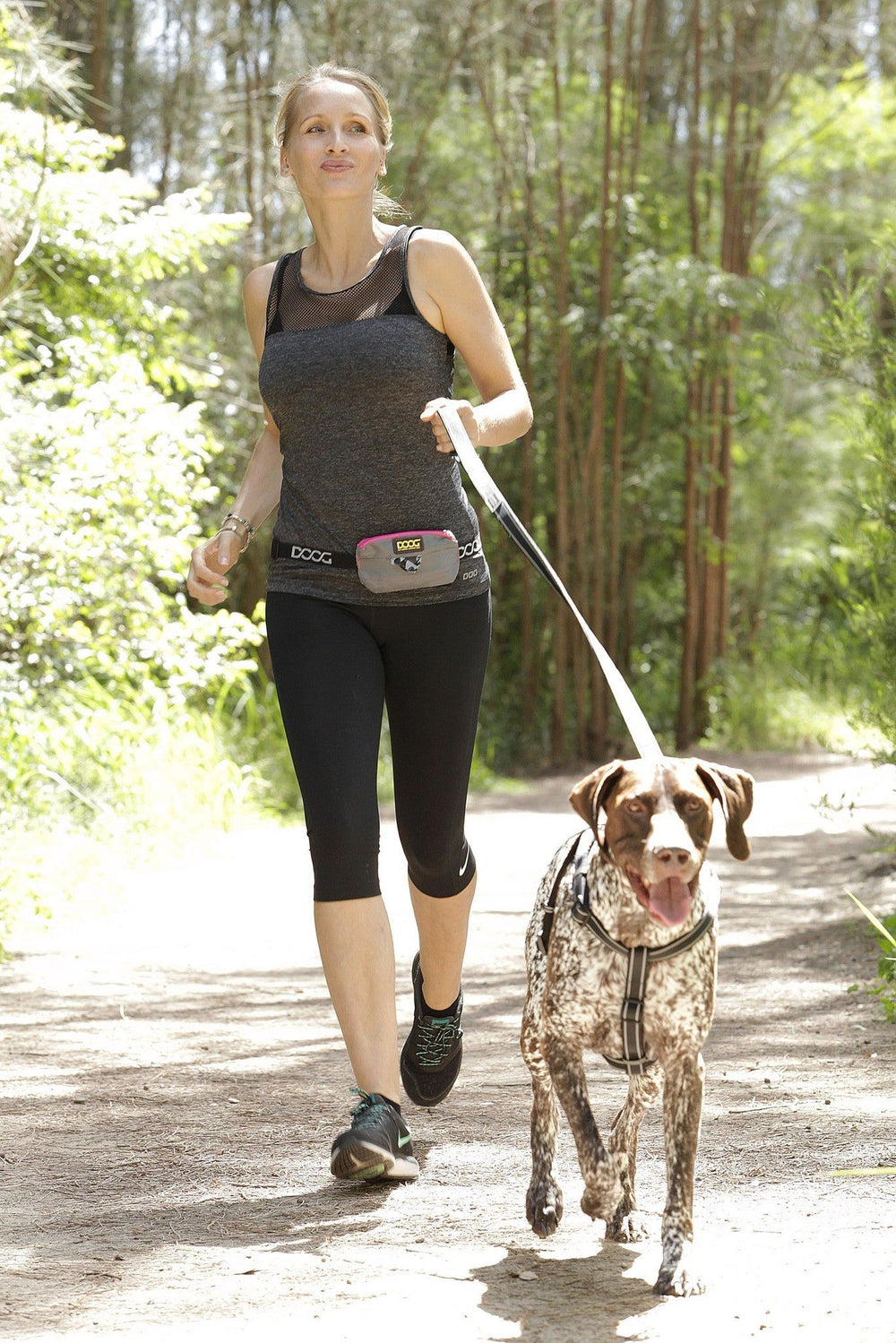 Frau mit Hund an der Leine, sportlich gekleidet in ärmellosem Oberteil und Leggings, fröhlicher Hund läuft.