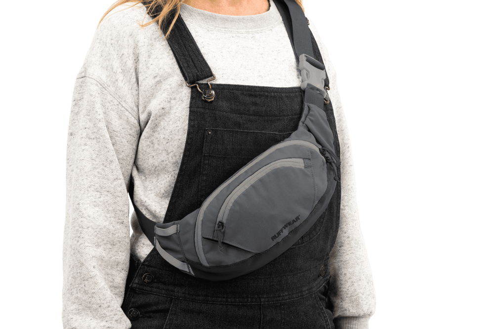 Person trägt graue Home Trail™ Hüfttasche über der Schulter, gekleidet in hellgrauen Pullover und schwarzen Latzhose.