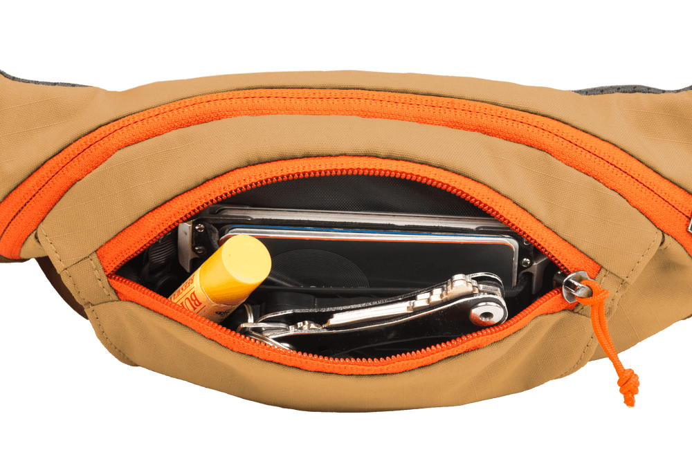 Beige Home Trail™ Hüfttasche mit orangefarbenen Reißverschlussdetails, gefüllt mit Smartphone, Lippenbalsam und Werkzeug.