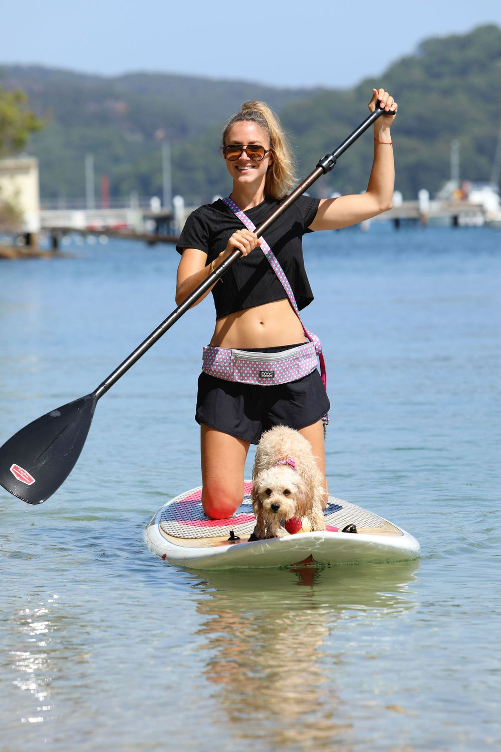 Junge Frau auf Stand-Up-Paddle-Board mit Hund, trägt DOOG Gürteltasche und Paddel im ruhigen Gewässer.