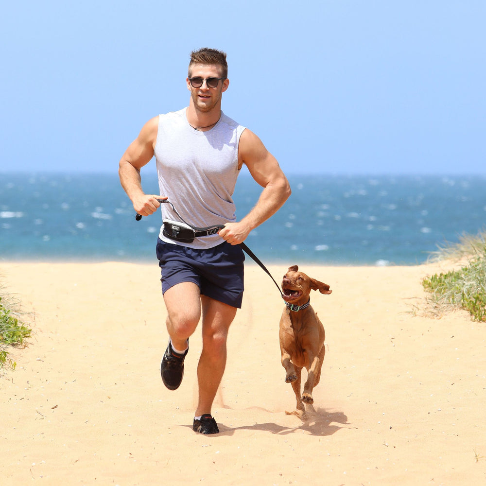 Muskulöser Mann joggt am Strand mit Hund an der Leine, trägt graues Tank-Top und Sonnenbrille.