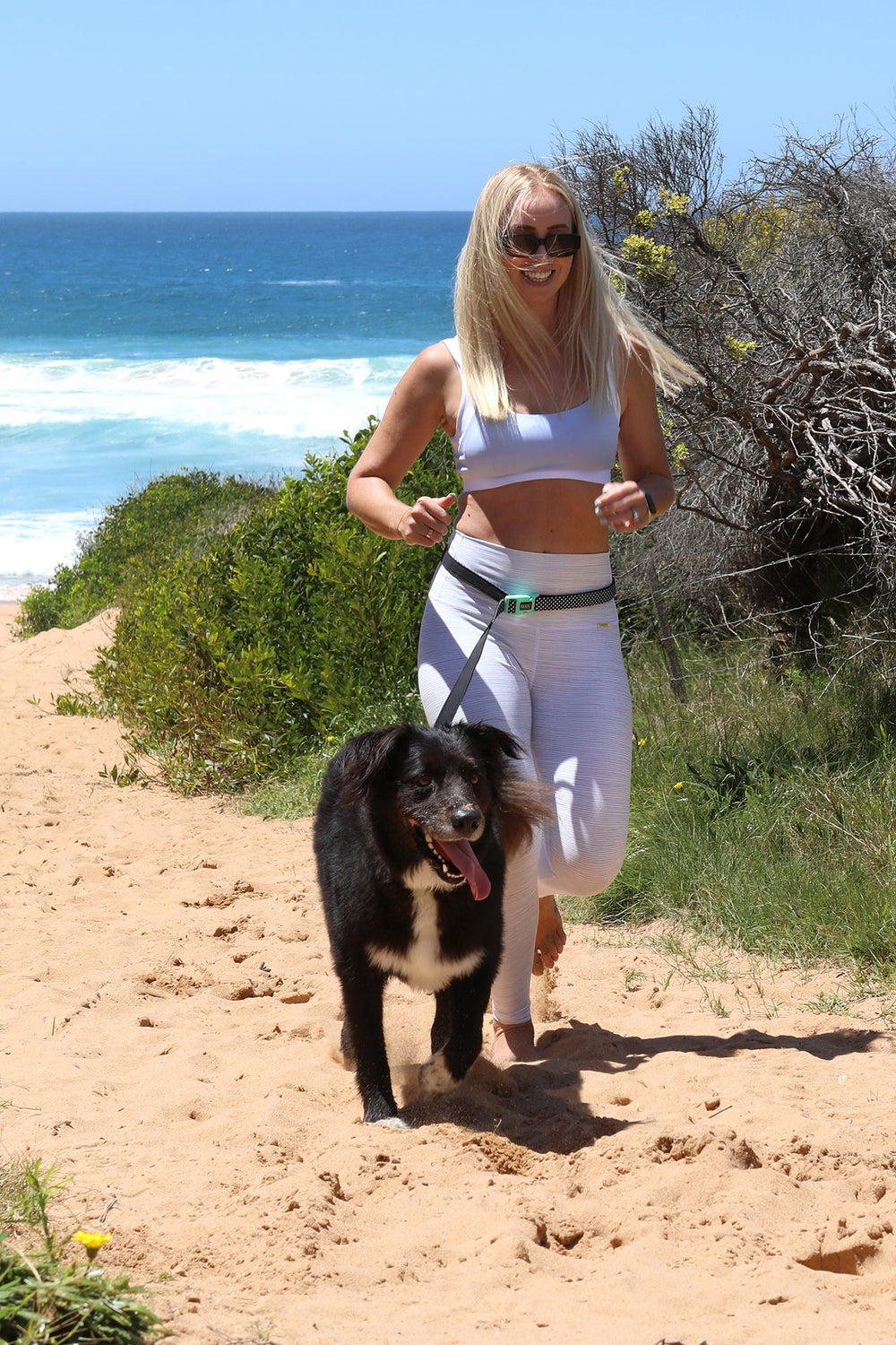 Frau mit Sonnenbrille spaziert freihändig am Strandweg mit schwarzem Hund unter blauem Himmel.