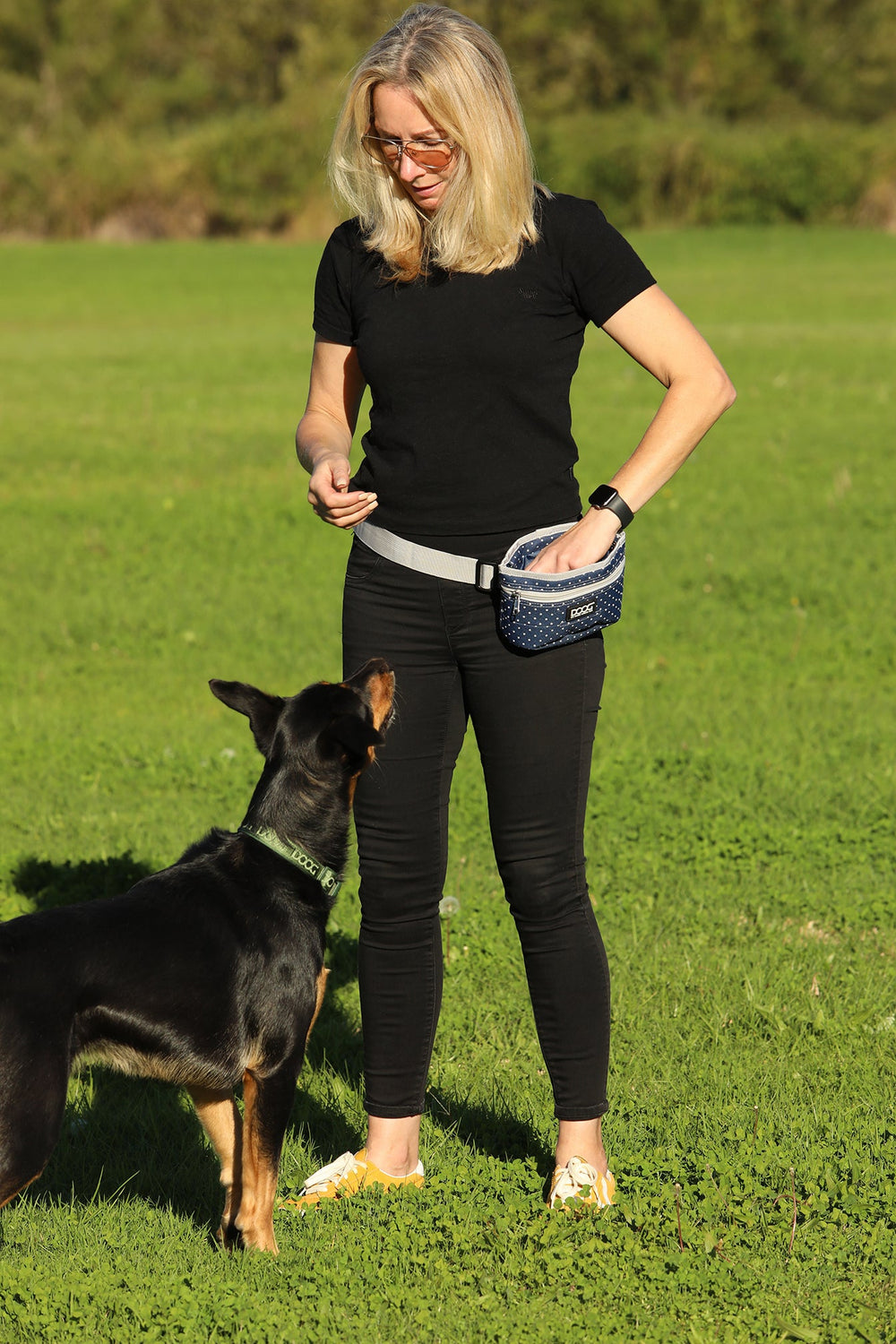 Blonde Frau mit Bauchtasche im grünen Feld, während sie einem Hund Leckerchen anbietet.