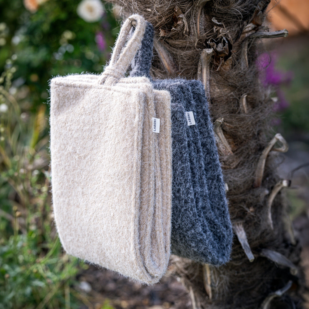 Zwei filzene Taschen in hellbeige und dunkelgrau hängen an einem Baumstamm, umgeben von blühenden Pflanzen.