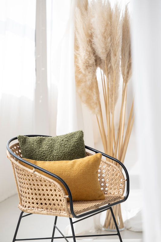 Moderne Rattanstuhl mit olivgrünem und senfgelbem Kissenbezug Snooze, umgeben von getrockneten Pampasgräsern.