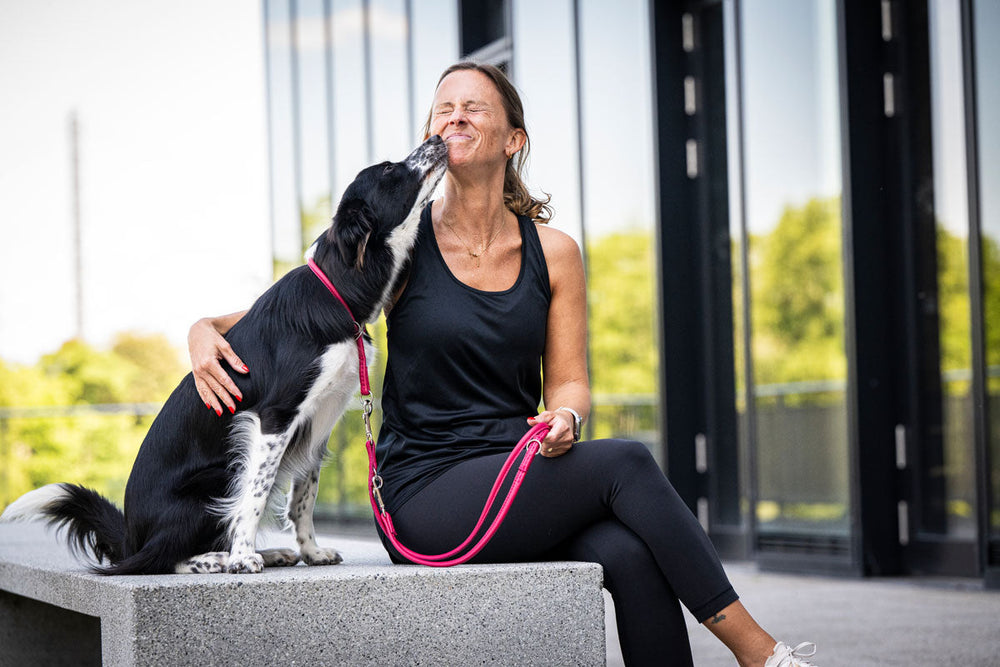 Frau in Sportkleidung mit Hund, der sie küssend anblickt, in heller, moderner Umgebung mit Glasflächen.