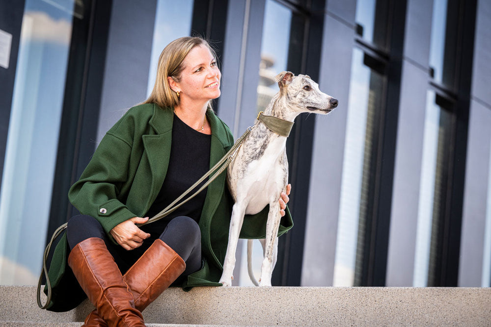 Frau in grünem Mantel mit grau-weißem Hund auf Stufen, handgelegt auf dem Hund, braune Stiefel sichtbar.