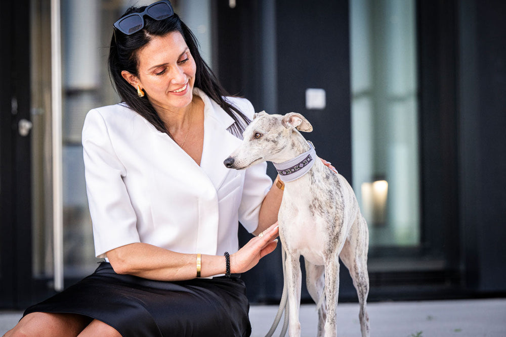 Eine Frau im weißen Blazer sitzt neben einem grau-weißen Windhund und lächelt freundlich.