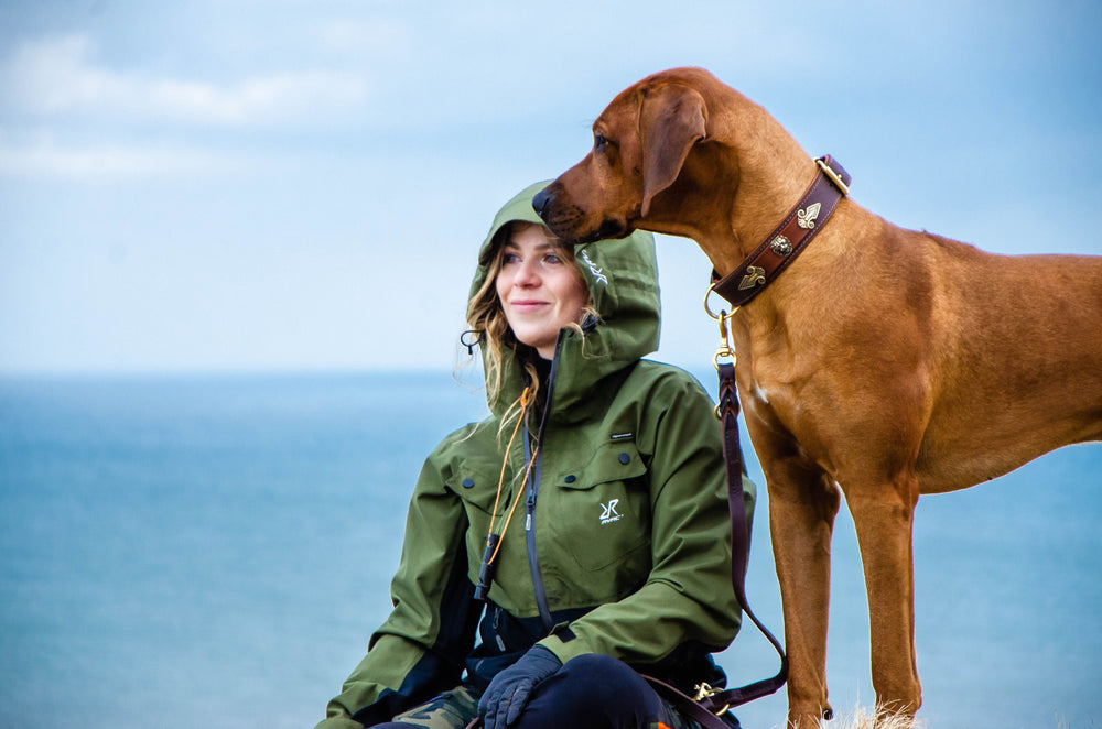 Frau in grüner Jacke mit großem Hund am Strand, Blick auf das Meer und bewölkten Himmel.
