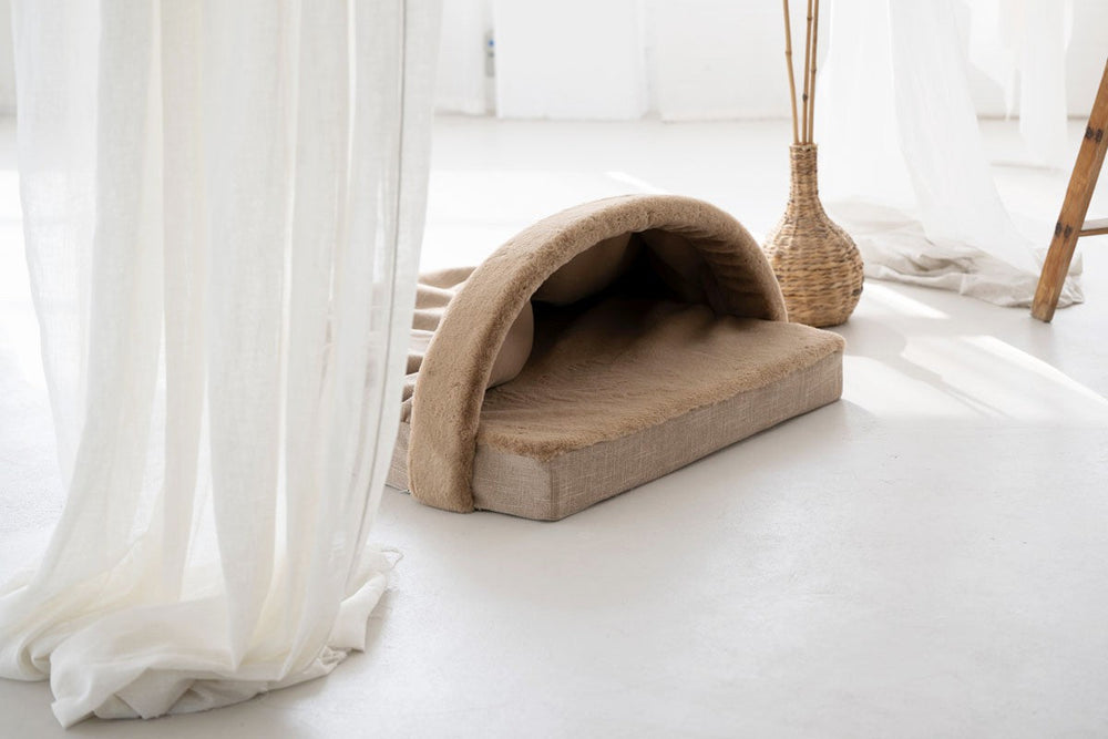 Gemütliches Hundebett in beige und braun mit Überhang, auf hellem Boden in einem lichtdurchfluteten Raum.