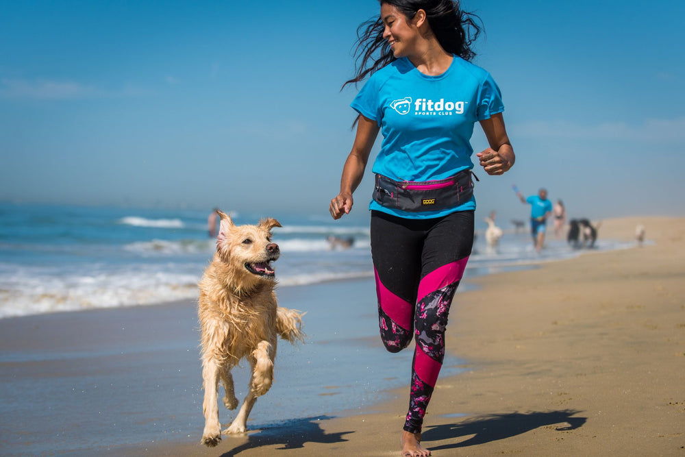 Joggende Frau am Strand mit goldenem Hund, sportliche Kleidung, blauer Himmel, fröhliche Atmosphäre und aktive Umgebung.