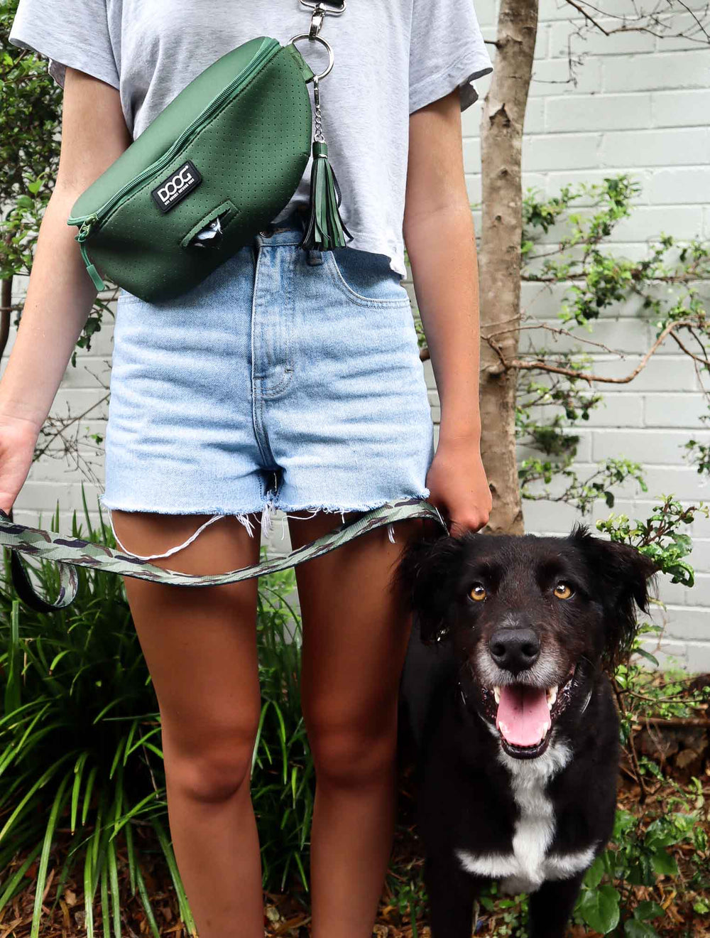 Person in Jeansshorts mit Umhängetasche und Hundeleine, begleitet von schwarzem Hund, trägt DOOG Neosport Hip Belt.