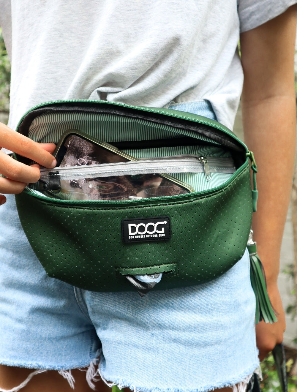 Grüne Umhängetasche geöffnet, innen transparente Tasche mit Dokumenten, Person in grauem T-Shirt und Jeansshorts.