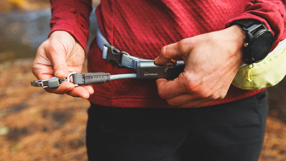 Person mit Hitch Hiker™ Leinensystem: Gürtel und Leine in der Hand, rotes Oberteil, schwarze Hose, unscharfer Hintergrund.