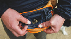 Person hält offene orangefarbene Home Trail™ Hüfttasche mit beige Zubehör, unscharfer Hintergrund zeigt weiteren Menschen oder Hund.
