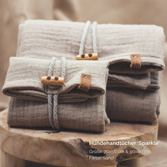 Zwei gefaltete Hundehandtücher in Sandfarbe mit dekorativer Schnur und Holzclip, Größen 70x50 cm und 90x60 cm.