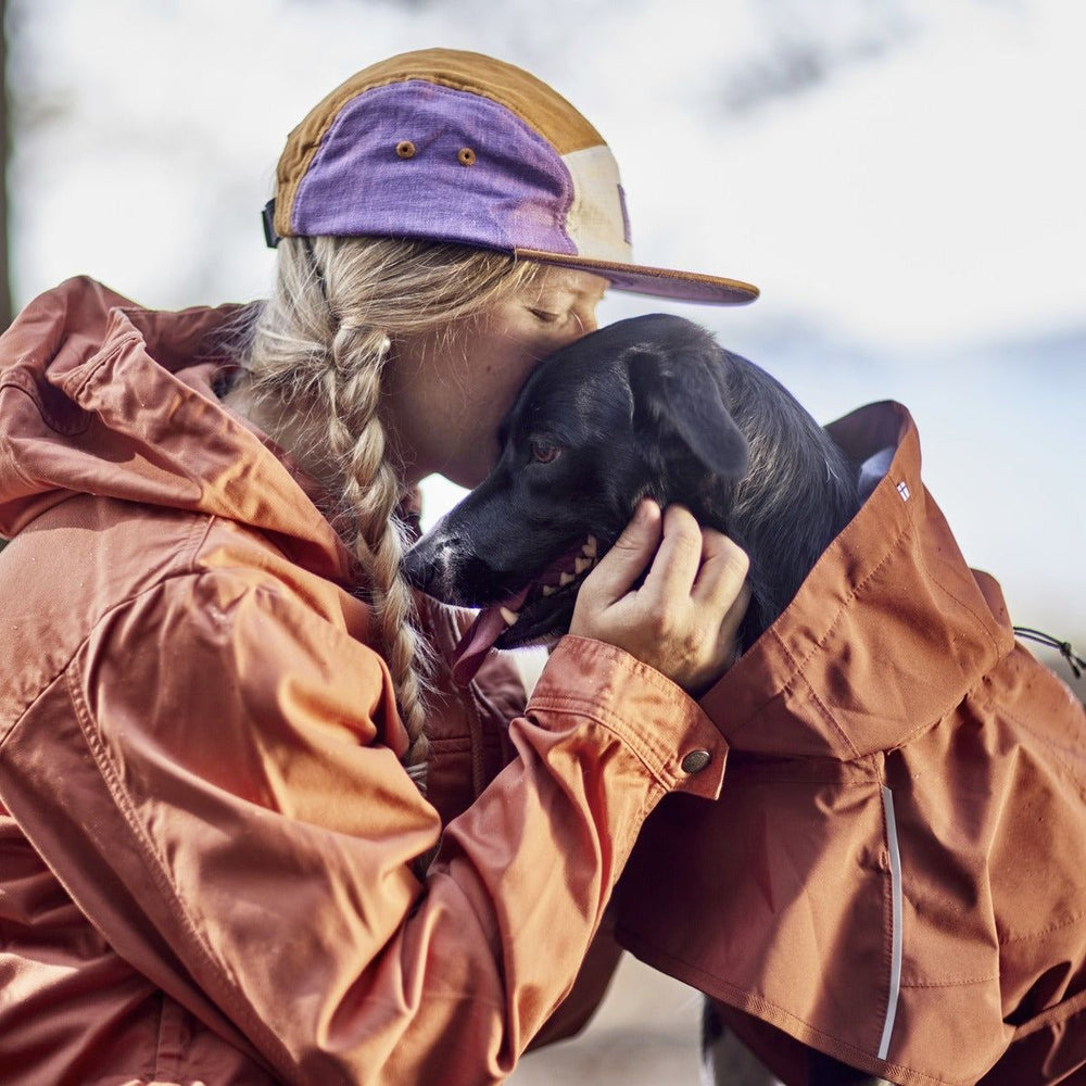 Person in orangefarbenem Regenmantel Mudventure Coat ECO umarmt schwarzen Hund in orangefarbener Jacke, beide mit bunter Kappe draußen.