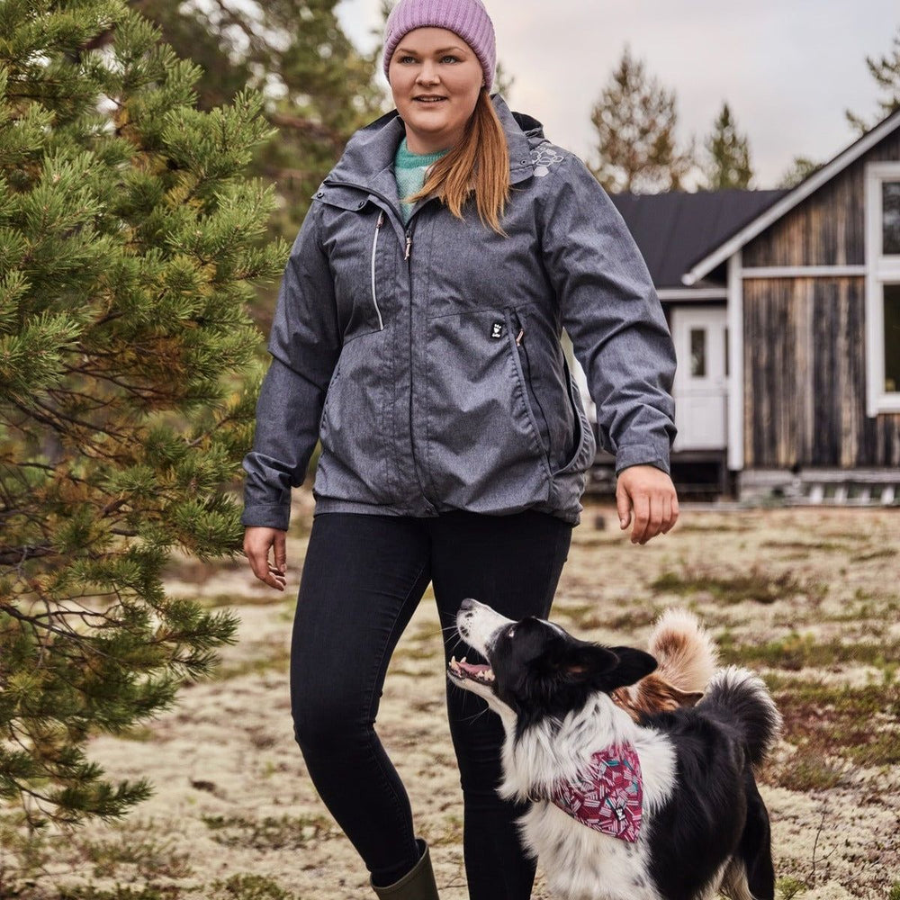 Graue ECO Trainingsjacke beim Spazieren mit Hund im Freien, kombiniert mit schwarzer Jeans und lila Mütze. Gebäude und Bäume im Hintergrund.