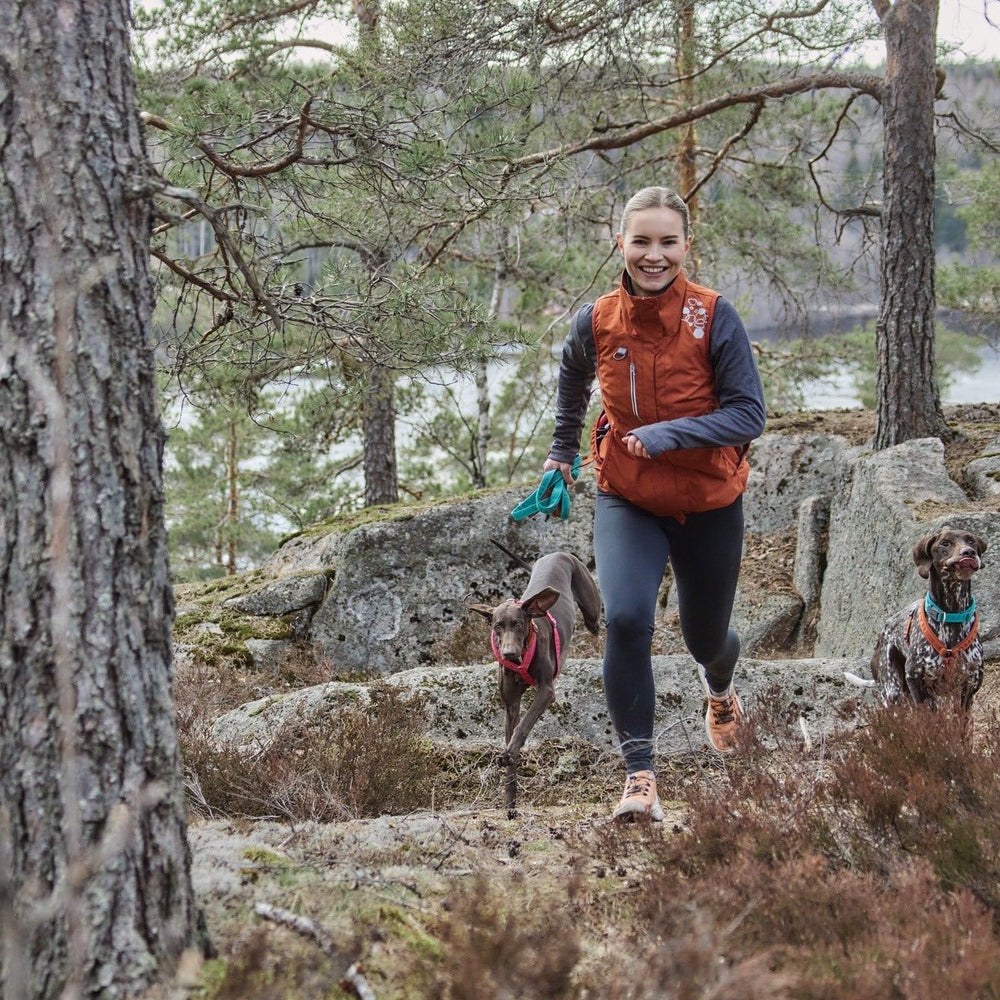 Joggerin im Wald mit Trainingsweste ECO, orange, und zwei Hunden an Leine; Hintergrund aus Bäumen und felsigem Gelände.