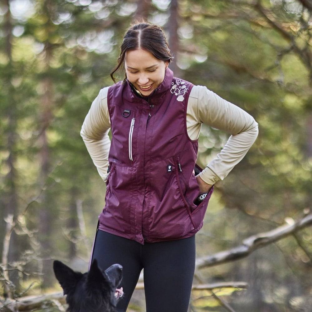 Frau in lila Trainingsweste interagiert lächelnd mit Hund im Wald zwischen Bäumen.