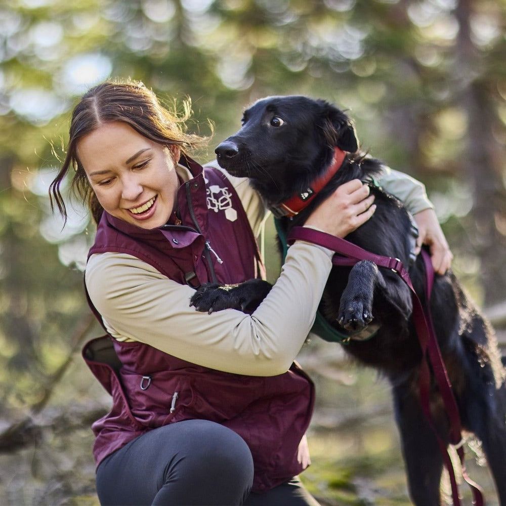 Junge Frau mit einem schwarzen Hund in den Armen, lächelnd in der Natur, trägt eine ärmellose Trainingsweste.