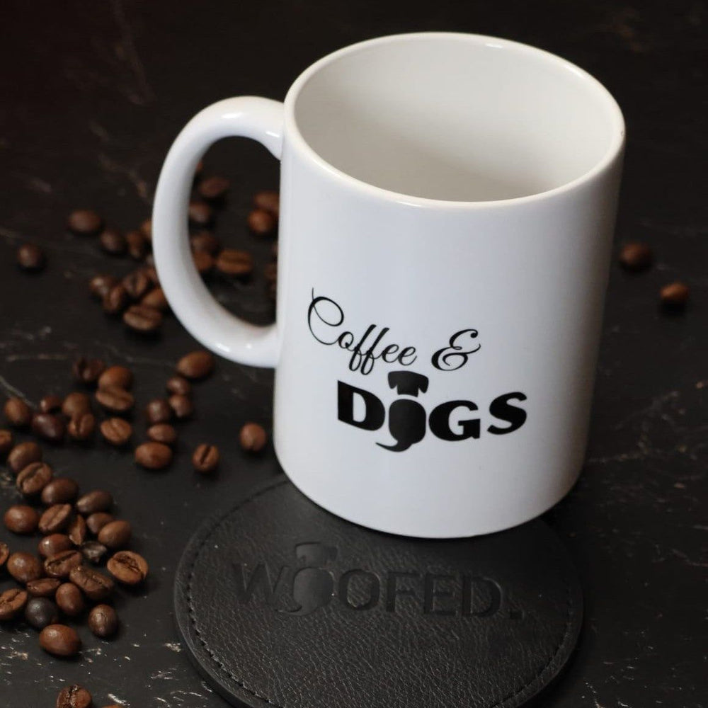Weiße Tasse mit "Coffee & Dogs" auf dunklem Untergrund, umgeben von Kaffeebohnen und einem Untersetzer mit "WOOF".