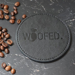 Runder schwarzer Leder-Getränkeuntersetzer mit „WOOFED“-Prägung, umgeben von Kaffeebohnen auf dunkler Oberfläche.
