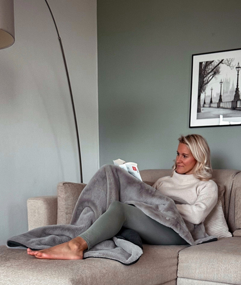 Person mit Webpelz-Wohndecke auf hellem Sofa, liest ein Buch in gemütlicher Atmosphäre.
