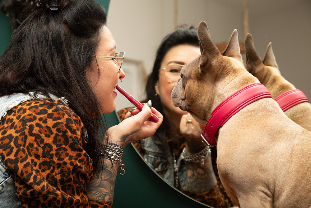 Frau schminkt sich im Spiegel, neben ihr sitzt ein aufmerksamer Hund mit rotem Halsband.