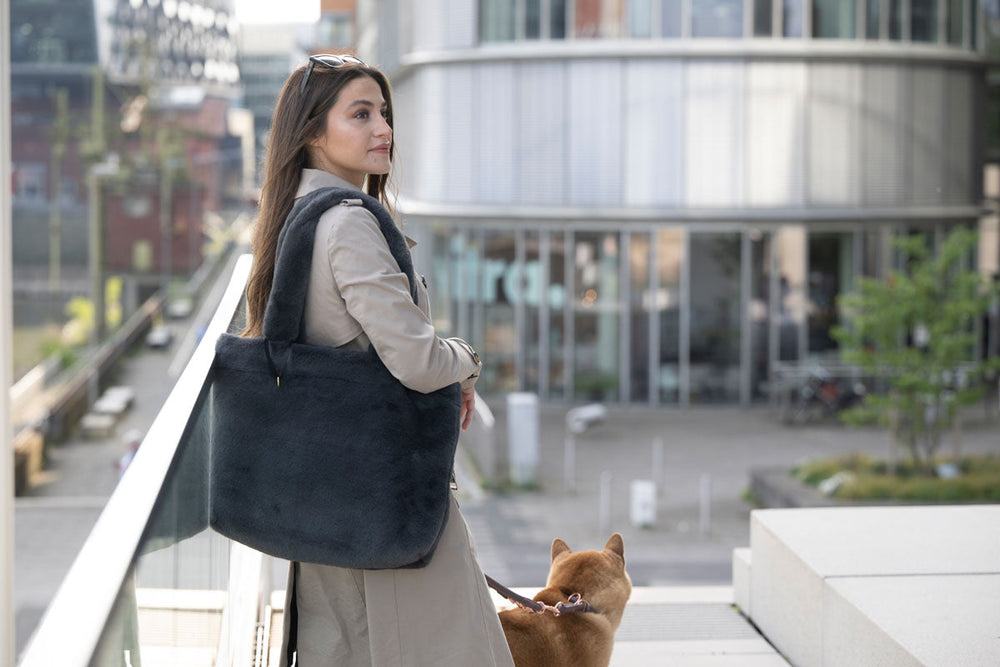 Frau mit braunen Haaren und beige Jacke auf einer Brücke, hält schwarze Pelztasche, neben ihr sitzt ein Hund.