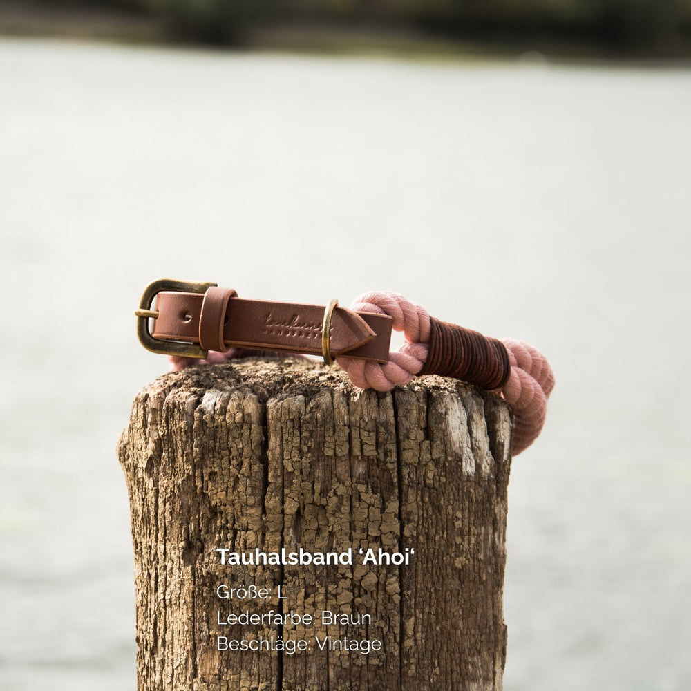 Braunes Tauhalsband "Ahoi" mit Vintage-Beschlag auf Holzpfahl, unscharfer Wasseroberfläche im Hintergrund.