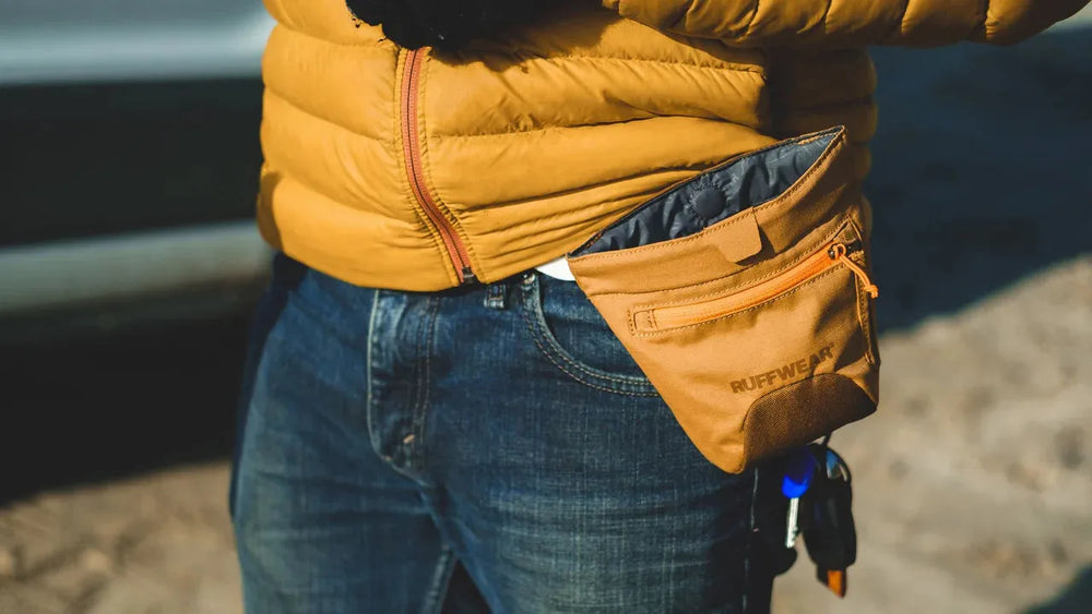 "Person in gelber Daunenjacke mit brauner Treat Trader™ Leckerlitasche am Gürtel und Jeans."