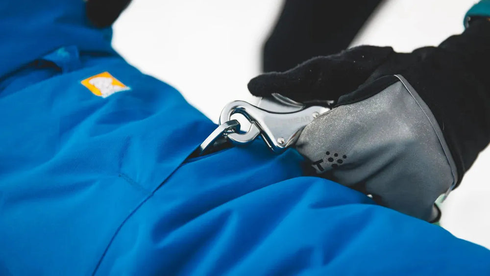"Hand mit schwarzem Handschuh befestigt Clip an blauer Vert™ Jacke vor schneebedecktem Hintergrund."