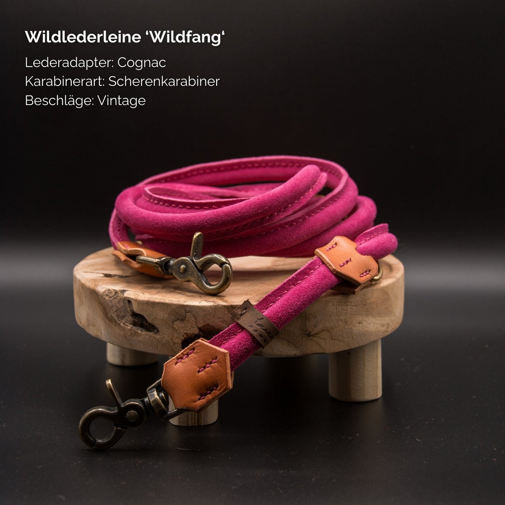 "Pinkfarbene Wildlederleine mit Cognac-Halter und Metallkarabiner auf Holzoberfläche drapiert."