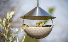 Hängender Vogelfutterspender in Schalenform mit gelb-grünen Vögeln beim Fressen, umgeben von unscharfem Grün.