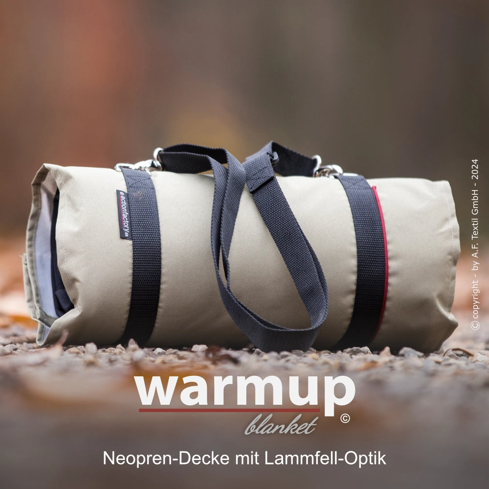 Beige Neopren-Decke in Lammfell-Optik, zusammengerollt mit roten Akzenten und Tragegriffen, auf unscharfem Hintergrund.