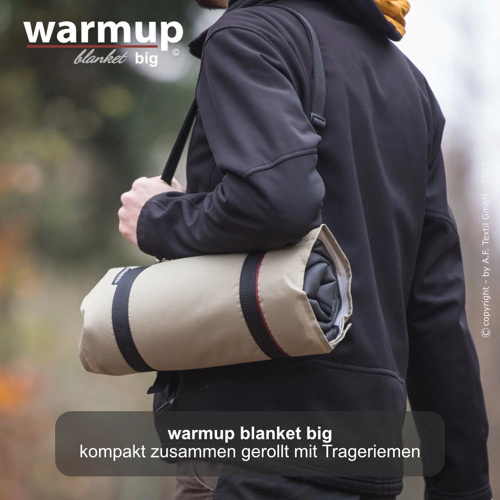 Person trägt eine kompakte, beige warmup© Decke mit Trageriemen über der Schulter, ideal für unterwegs.