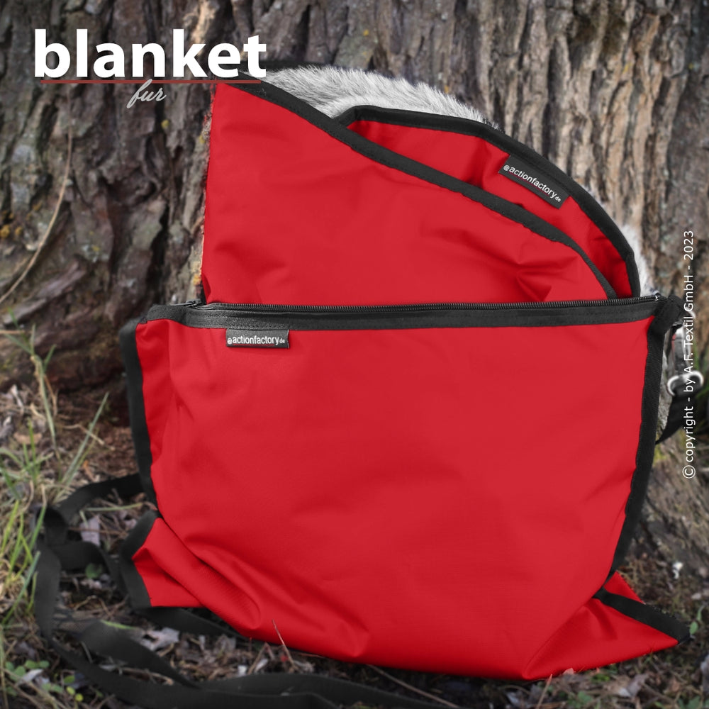 Rote Decke mit grauem Innenfutter, zusammengefaltet und von schwarzer Umrandung gehalten, vor einem Baumstamm.