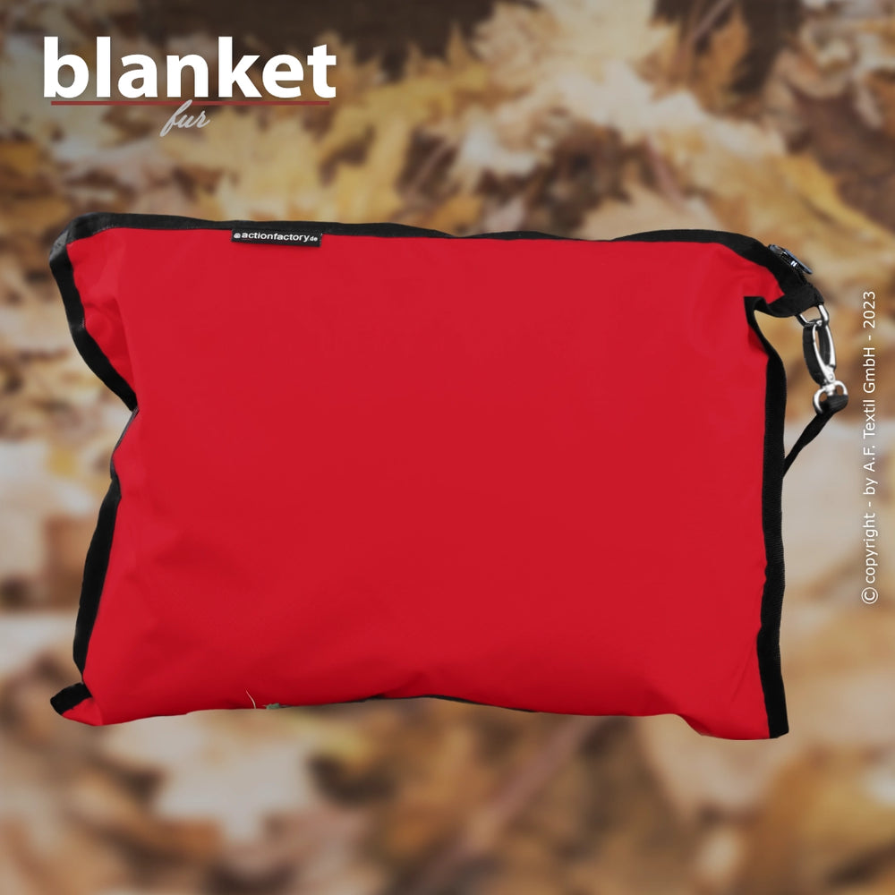 Rotes Kissen mit schwarzem Reißverschluss und Karabiner vor unscharfem, herbstlichem Blätterhintergrund.