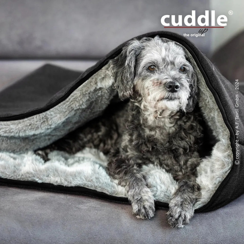 Grauer Hund kuschelt sich in eine schwarze und helle Decke auf einem bequemen Hundeliegeplatz von cuddle.up®.