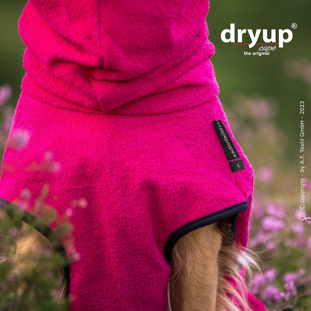 Hund trägt pinken Fleece-Bademantel mit Kapuze, umgeben von blühenden Pflanzen, ideal für nasse Hunde.