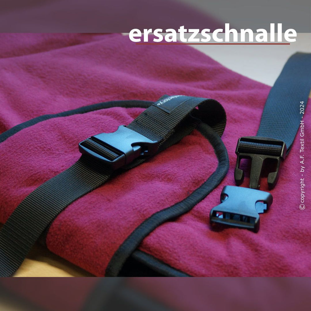 Ersatzschnalle aus Kunststoff neben schwarzem Gurt auf rotem, weichem Untergrund, der obere Teil abgenommen.