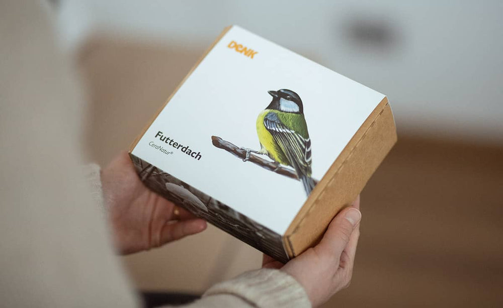 Person hält eine Box mit dem Produkt "Futterdach" für Meisenknödel, dekoriert mit einem bunten Vogel.